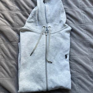 Ljusgrå hoodie från Polo Ralph Lauren - Snygg ljusgrå hoodie från Polo Ralph Lauren med dragkedja, huva och snörning. Klassisk logga broderad på bröstet och praktiska fickor framtill. Perfekt för en chill och stilren look.