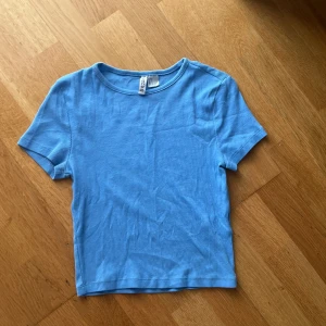 Blå ribbad topp från H&M Basic - En enkel och snygg blå ribbad topp från H&M Basic. Modellen är kortärmad med rund halsringning och har en tight passform. Perfekt att matcha med jeans eller kjol för en clean look.