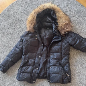 Svart pufferjacka med äkta päls från Rockandblue - Säljer en svart pufferjacka från Rockandblue med snyggt quiltat mönster och stor huva med brun äkta päls. Jackan har dragkedja, knappar och två fickor med dragkedja framtill. Perfekt för kalla dagar och riktigt skön att bära. pris kan diskuteras