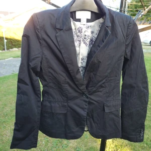Svart kavaj från H&M, storlek 36 - Stilren svart kavaj från H&M i storlek 36. Klassisk modell med krage, knappstängning och två fickor framtill. Fodret har ett diskret blommigt mönster i vitt och lila.