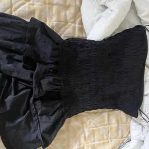 Svart axelbandslös kort klänning - Säljer en svart axelbandslös kort klänning med smockad överdel och volangig kjol. Klänningen har en snygg och trendig look, perfekt för dig som vill sticka ut. Materialet känns mjukt och följsamt mot kroppen.