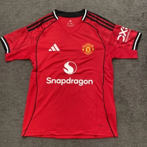 Manchester United Adidas fotbollströja M - Röd Manchester United fotbollströja från Adidas med klubbmärke, Snapdragon-sponsor och svarta detaljer. Klassisk V-ringning, svarta ränder på axlarna och texten 'Theatre of Dreams' i nacken. Tillverkad i lätt och ventilerande polyester.
