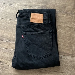Svarta Levi's 501 straight jeans - Svarta Levi's 501 jeans med klassisk rak passform och fem fickor. Jeansen har knappgylf, snygga nitar och den ikoniska läderpatchen bak i midjan. Tillverkade i robust denim som ger en tidlös look. Perfekta för dig som gillar en clean och stilren vibe.