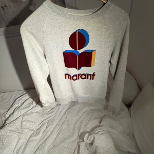 Grå sweatshirt från Isabel Marant - Grå sweatshirt från Isabel Marant med ikoniskt tryck i vinrött, blått och gult framtill. Tröjan har rund hals, långa ärmar och ribbade muddar. Perfekt för en chill och snygg look.