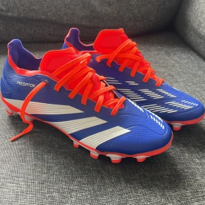 Adidas Predator blå fotbollsskor 41⅓ - Säljer ett par Adidas Predator fotbollsskor i blått med vita detaljer och orange snörning. Skorna har en syntetisk ovandel med Hybridfeel-textur och mönstrad sula för bra grepp på planen. Perfekta för dig som vill sticka ut på matchen!