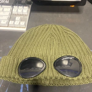 Olivgrön goggle-mössa från C.P. Company - Säljer en grön cp company mössa med dom kända glasögonen på 