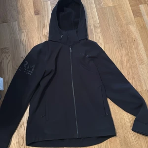 Svart vindjacka från Race Marine - Svart vindjacka från Race Marine med huva och hel dragkedja framtill. Jackan har diskret tryck på ena ärmen och två fickor med dragkedja. Snygg och enkel design som passar perfekt till vardags. Materialet känns mjukt och smidigt.