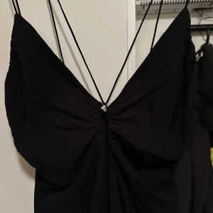 Svart långklänning från H&M strl: XS - Svart långklänning från H&M med smala axelband och snygg rynkad detalj framtill. Klänningen har djup v-ringning och dragkedja i sidan för enkel påklädning. Perfekt för dig som gillar stilrena och eleganta plagg.