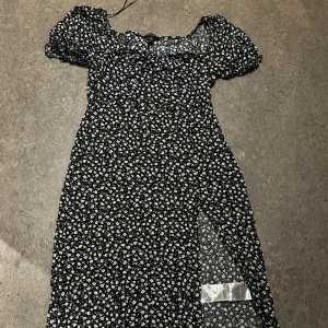 Svart blommig klänning med slits - Supersöt svart klänning med små vita blommor, puffiga korta ärmar och en snygg slits framtill. Klänningen har en smickrande passform och offshoulder-axlar som ger en trendig vibe. Den är pyttelite sönder under armen vid dragkedjan men det är lätt att fixa eller sy själv. Säljer för 40kr