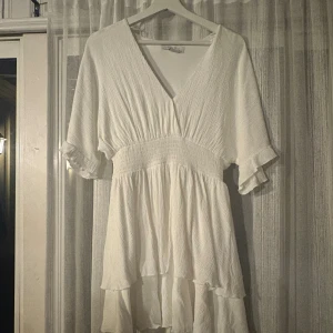 Vit omlottklänning från Amisu - Superfin vit kortklänning från Amisu med v-ringning, omlottkänsla och volangdetaljer. Klänningen har korta ärmar med lätt rynk och smockad midja som ger en snygg siluett. Perfekt för sommarens alla tillfällen! Endast använd en dag på skolavslutning