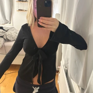 Svart omlottopp med knyt - Säljer en svart omlottopp från Gina med lång ärm och djup v-ringning. Toppen har en snygg knytning framtill som ger en cool och trendig look.