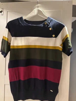 Randig stickad topp från Holly & Whyte - Säljer en snygg randig topp från Holly & Whyte i storlek M. Toppen har breda horisontella ränder i marinblått, vitt, olivgrönt, vinrött och gult. Rund halsringning med tre dekorativa guldfärgade knappar på axeln. Kortärmad och mjukt stickad material.