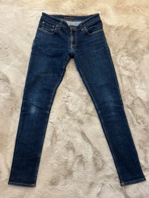  Nudie Jeans - Säljer ett par mörkblå jeans från Nudie Jeans med klassisk femficksdesign och orangea kontrastsömmar. Jeansen har låg midja och är tillverkade i stretchig bomull för en tight passform. Perfekt för dig som gillar en smal siluett och stilrena detaljer, jeansen har dessutom slitningar perfekt för dig som gillar stil och mode. Kom dm för eventuella frågor 🧐pris kan diskuteras 💯😇