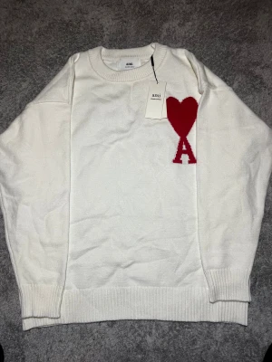 Vit stickad tröja från Ami - Vit stickad tröja från Ami med rund hals och ribbade muddar. På bröstet finns ett stort rött hjärta och bokstaven A som snygg detalj. Perfekt för dig som gillar stilrena plagg med en twist. Materialet är mjukt och skönt, passar till många olika looks.