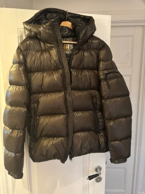 Svart pufferjacka från Hetrego - Säljer en grön pufferjacka från Hetrego med huva och stora quiltade partier. Jackan har dragkedja framtill, två sidofickor med dragkedja och en extra ficka på ärmen.  Jackan är använd i en vecka så väldigt bra skick. Storlek S. Modellen heter Scott nypris 4900kr. 
