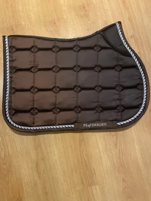 Brunt schabrak från PS of Sweden - Säljer ett brunt schabrak från PS of Sweden i storlek COB.  schabraket har snygga broderade detaljer och en vit tvinnad kant. Materialet är mjukt och glansigt, perfekt för dig som gillar stilren utrustning till ridningen. Texten 'Ride more, worry less' finns broderad på sidan.