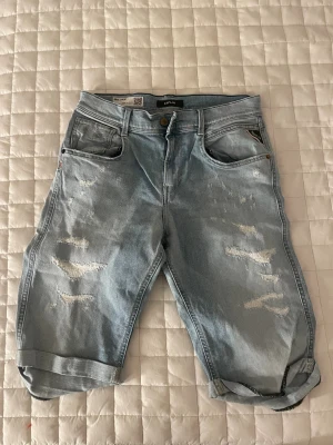 Replay ljusblå jeansshorts med slitningar - Säljer ett par ljusblå jeansshorts från Replay med snygga slitningar och uppvikta benslut. Klassisk femficksmodell med knapp och dragkedja framtill samt Replay-logga på bakfickan. Perfekt för dig som gillar en avslappnad och trendig stil.