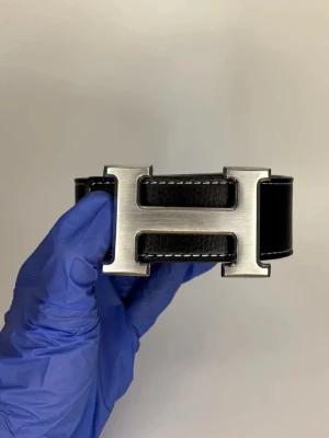 Svart skinnväst med metallspänne - Snygg svart väst i skinn med ett stort, stilrent metallspänne framtill i form av ett H. Västen har en klassisk design med markerade sömmar och ger en cool och modern vibe. Perfekt för dig som vill sticka ut med en unik detalj.