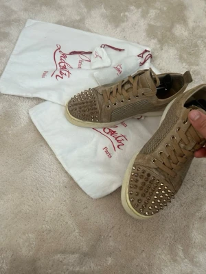 Beige Christian Louboutin sneakers med nitar - Säljer ett par beige sneakers från Christian Louboutin med klassisk röd sula och coola metallnitar på tån. Skorna har snörning, rund tå och är tillverkade i mocka och mesh. Perfekt för dig som vill sticka ut med exklusiva detaljer och lyxig känsla. Det går att göra dem renare!!! Pris går att diskuteras vid snabb affär