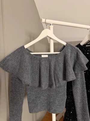 Grå stickad tröja med volang från H&M - Säljer en grå stickad tröja från H&M i storlek XS. Tröjan har en bred volang över axlarna och långärmade ärmar. Används ej längre🤍
