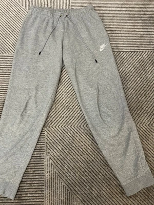 Grå mjukisbyxor från Nike, storlek M - Säljer ett par grå mjukisbyxor från Nike i storlek M. Byxorna har elastisk midja med snörning och ribbade muddar vid bensluten. Materialet är mjukt och skönt, perfekt för chill dag. 
