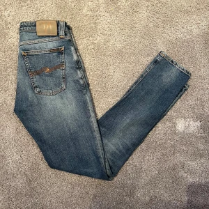   Nudie jeans - Säljer ett par blå  jeans från Nudie Jeans med klassisk femficksdesign och snygga kontrastsömmar. Jeansen har en mörk tvätt med  och detaljerade bakfickor med Nudie-loggan. Perfekt passform och bekväm stretch i materialet. Storleken är 30 32 och de är i nytt skick!