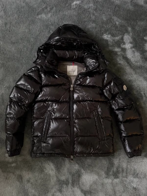 Moncler Maya  - Säljer en svart pufferjacka från Moncler med glansig finish och huva. Jackan har dragkedja framtill, sidofickor med dragkedja och Moncler-logga på ärmen. Insidan har ett snyggt serietryck och nederkanten är justerbar. Perfekt för det kallare vädret som är på väg ⛄️❄️