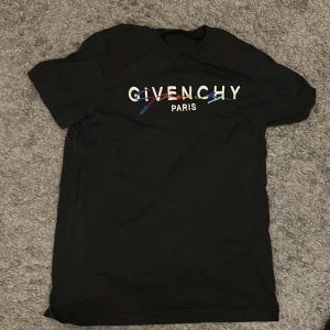 Svart Givenchy t-shirt med färgglatt tryck - Säljer en svart t-shirt från Givenchy med ett stort, vitt tryck på bröstet där det står 'GIVENCHY PARIS' och en färgglad grafisk linje över texten. T-shirten har rund hals och korta ärmar, tillverkad i mjuk bomull för skön känsla.
