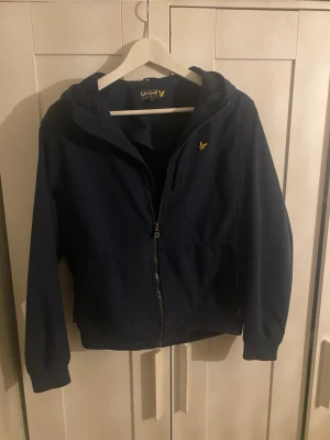 Marinblå vindjacka från Lyle & Scott - Snygg marinblå vindjacka från Lyle & Scott med huva och gul logga på bröstet. Jackan har dragkedja framtill, ribbade muddar och är tillverkad i polyester. Perfekt för dig som vill ha en stilren och enkel jacka med skön passform.