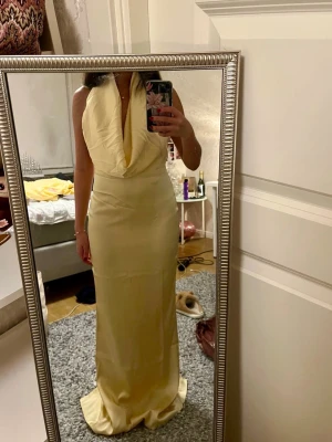 Gul  långklänning i satin - Balklänning i satin med halterneck och draperad urringning. Klänningen har öppen rygg, lång knytbanddetalj och slits bak.                                             Helt ny med alla lappar kvar, returtiden är ute. Storlek 6 men skulle klassa som en 34/32