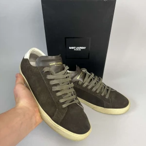 Bruna Saint Laurent sneakers i mocka - Snygga bruna sneakers från Saint Laurent Paris i mjuk mocka med klassisk låg profil. Skorna har grå snörning, vit sula och detaljer i läder på plösen med guldtext. Perfekta för dig som gillar stilrena och exklusiva sneakers. Pris kan diskuteras vid snabb affär. OBS Paket medföljer inte, därav låga priset