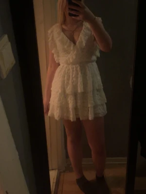 Vit volangklänning med spetsdetaljer,   - Säljer en söt vit kortklänning med volanger och spetsdetaljer. Klänningen har omlott-v-ringning, är ärmlös och har en öppen rygg med knyt i nacken. Materialet är lätt och perfekt för varma dagar.  Använd 1 gång och köpte klänningen för 600kr så säljer den för 400 kr,storlek S 