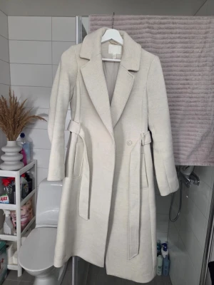 Beige kappa från H&M med bälte - Säljer en stilren beige kappa från H&M i storlek 32. Den har klassisk krage, långa ärmar och ett matchande bälte i midjan. Kappan är fodrad och har en enkel knappstängning framtill. Perfekt för dig som gillar minimalistisk och clean look. 