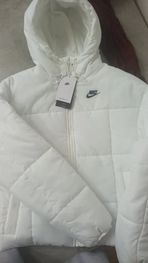 Vit pufferjacka från Nike Therma-Fit XS - Säljer en fräsch vit pufferjacka från Nike Therma-Fit i storlek XS. Jackan har huva med snörning, dragkedja framtill och två sidofickor. Perfekt för kalla dagar och har en clean, sportig look med Nike-logga på bröstet.