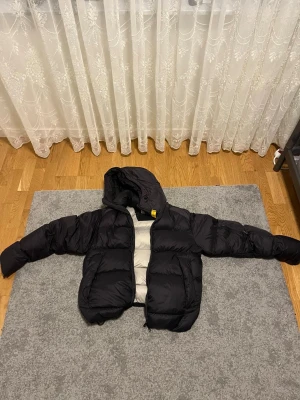 Parajumper tyrik - Svart dunjacka från Parajumpers med huva och coolt gult spänne vid halsen. Jackan har flera fickor, dragkedja framtill och patch på ärmen. Insidan är ljusgrå och materialet är polyester. Perfekt för kalla dagar och riktigt snygg passform.