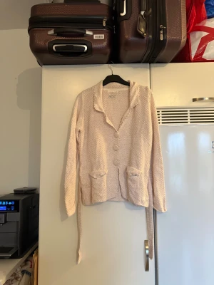 Beige stickad kofta från 506 - Säljer en beige och ljusrosa stickad kofta med grovstickad struktur, stora knappar framtill och två fickor. Koftan har lång ärm, krage och ett bälte i midjan för extra stil. Perfekt för lager-på-lager under kyliga dagar.  Storlek 2 som enligt deras storleksguide motsvarar en M.  Mycket fint skick, jag hittar inga synliga anmärkningar eller defekter. 