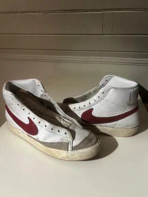 Nike Blazer Mid vit/röd (UTAN SKOSNÖRE) - Nike Blazer Mid sneakers i vitt skinn med röd swoosh på sidorna. Klassisk retrodesign med högt skaft och platt sula. Grå mockadetaljer vid tån och perforeringar för snörning. Yttersulan har fiskbensmönster för extra grepp. (UTAN SKOSNÖRE)
