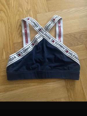 Mörkblå linnetopp Tommy Hilfiger - Säljer en mörkblå linnetopp från Tommy Hilfiger i storlek M. Toppen har breda vita axelband med logotyp och korsad rygg. Materialet är mjukt och stretchigt, perfekt för en sportig look. Snygg branding längs banden och klassisk design.