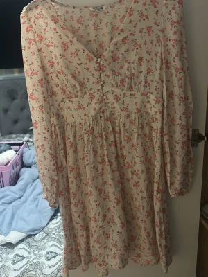 Blommig kortklänning från VILA - Supersöt kortklänning från VILA i ljus beige med små rosa blommor. Klänningen har v-ringning, knappar framtill och långärmade ärmar med resår vid handlederna. Smockad rygg ger en skön passform och det tunna materialet känns luftigt och fräscht.