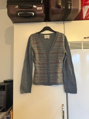 Grå kofta med mönster från Fira - Supermysig grå kofta från Fira med färgglada zigzag-mönster i orange, lila och vitt. Koftan har v-ringning, knappar framtill och långa ärmar. Perfekt att slänga på sig när det är kyligt ute! Mycket fint skick, men ytludd och noppror kan förekomma eftersom materialet är ull blandning. Storlek M