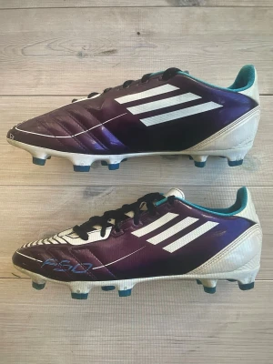 Adidas F50 lila/vita fotbollsskor - Säljer ett par Adidas F50 fotbollsskor i lila och vitt med klassiska tre vita ränder på sidan. Skorna har svarta snören, blå detaljer och en låg profil med dobbar för gräsplan. Insidan är turkos och ovandelen är i syntetmaterial för lätt vikt och bra bollkontroll.