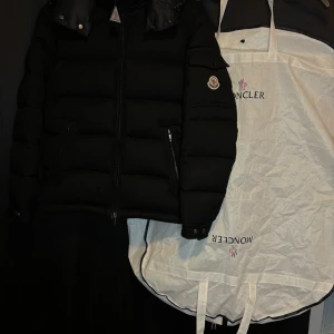 Svart pufferjacka Moncler - Svart pufferjacka från Moncler med huva och dragkedja framtill. Jackan har Moncler-logga på ärmen, två sidofickor med dragkedja och quiltad design. Perfekt för kalla dagar och har en stilren look som funkar till många outfits.