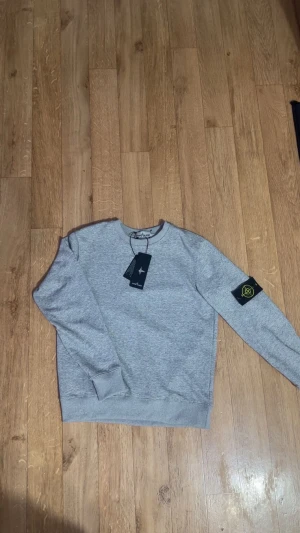 Grå sweatshirt från Stone Island - Snygg grå sweatshirt från Stone Island med klassisk rund hals och ribbade muddar. Tröjan har den ikoniska Stone Island-patchen på vänster ärm och är tillverkad i mjuk bomull. Perfekt för dig som gillar stilrena och bekväma plagg.