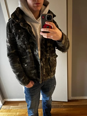 Camouflage parkajacka från Black - Säljer en grön och brun camouflage fieldjacket från Black med flera stora fickor och snygga metalldetaljer. Jackan har dragkedja och knappar framtill, hög krage och cool militärstil. Perfekt för dig som vill sticka ut med streetwear-vibe. Skriv vid funderingar😁