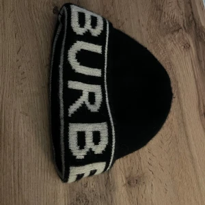 Svart Burberry mössa i kashmir - Svart mössa från Burberry med vit logotyp runt kanten. Tillverkad i 100% kashmir för extra mjuk och lyxig känsla. Klassisk form och stilren design som sticker ut med den tydliga Burberry-texten. Perfekt för dig som gillar exklusiva accessoarer.