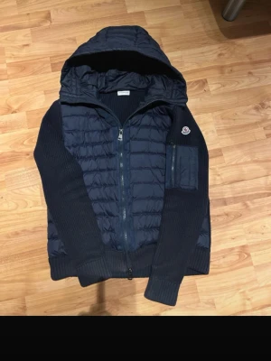 Mörkblå Moncler Cardigan NFC Scan  - Storlek L fast passar M. Snygg mörkblå hybridjacka från Moncler med quiltad front och huva. Jackan har stickade ärmar och sidor, dragkedja framtill och en praktisk ficka med dragkedja på ärmen. Perfekt för dig som vill ha en sportig och stilren look.