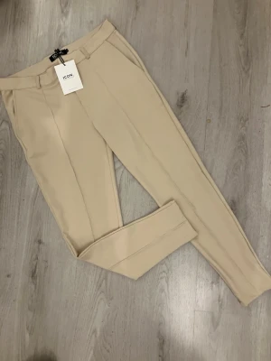 Beige kostymbyxor från ICON Amsterdam - Snygga beige kostymbyxor från ICON Amsterdam i storlek L. Byxorna har raka ben, pressveck och bälteshällor. De är tillverkade i ett mjukt material som ger en clean och stilren look. 