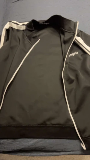 Svart Palm Angels M - Svart vindjacka från Palm Angels med vit dragkedja och vita ränder längs axlarna. Jackan har ståkrage och logotypen tryckt på bröstet. Tillverkad i polyester för en sportig och clean look. Perfekt för dig som gillar streetwear och vill sticka ut.