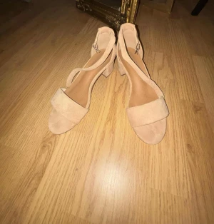 Beige sandaler med klack - Säljer ett par snygga beige sandaler med bred rem över tårna och öppen tå. Skorna har en stabil blockklack och är gjorda i mocka. Perfekta för varma dagar och passar till många olika outfits.
