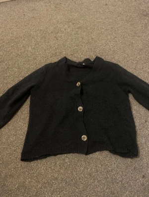 Mörkblå kofta med knappar - Mörkblå kofta med v-ringad hals och tre stora knappar framtill. Långärmad modell med ribbade detaljer vid midjan och ärmslut. Perfekt för lager-på-lager och enkel att matcha med olika outfits.